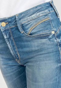Jeans in denim azzurro chiaro presentano una vestibilità slim, cuciture visibili, una tasca per le monete e accessori in tonalità oro sulla vita anteriore.