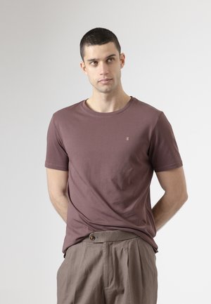 Joven con cabello corto y oscuro que lleva una camiseta malva y pantalones plisados beige, de pie con las manos detrás de la espalda, mirando hacia la derecha.