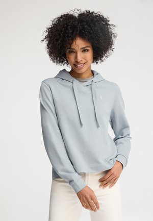 Femme aux cheveux bouclés portant un sweat à capuche bleu clair et un pantalon blanc, souriante, les mains légèrement jointes à la taille.