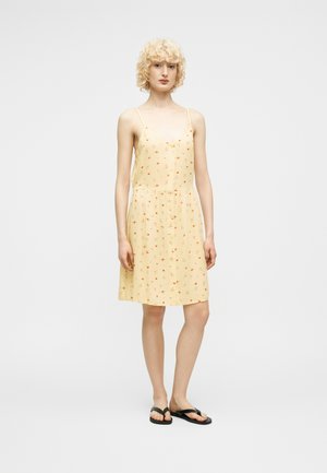 Młoda osoba o kręconych blond włosach, ubrana w żółtą, bezrękawnikową sukienkę z małym kwiecistym wzorem oraz czarne japonki, stojąca na jednolitym tle.