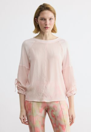 Νεαρή γυναίκα με κοντά ξανθά μαλλιά φοράει ανοιχτό ροζ μπλούζα με μακριά μανίκια και ρυτιδωτά μανίκια, καθώς και παντελόνια σε παστέλ πολύχρωμα, στέκεται μπροστά σε λευκό φόντο.