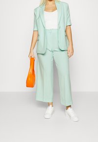 Muntgroene blazer en wijde broek gecombineerd met een witte top en oranje handtas; witte sneakers maken de outfit compleet. Gladde textuur.