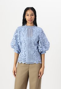 Lyseblå blondebluse med puffy ermer, intrikate blomster mønstre, h�øy hals og bølget falm, kombinert med beige bukser.