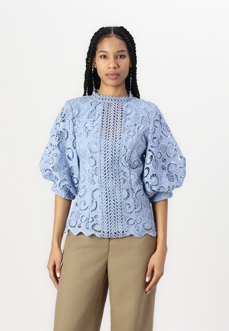 Lyseblå blondebluse med puffy ermer, intrikate blomster mønstre, høy hals og bølget falm, kombinert med beige bukser.