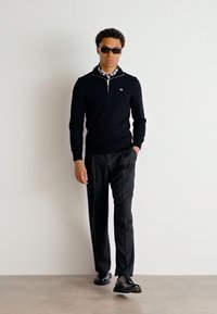 Pulover negru cu guler cu fermoar peste o cămașă cu carouri, asortat cu pantaloni negri și pantofi negri. Modelul poartă ochelari de soare închiși la culoare.