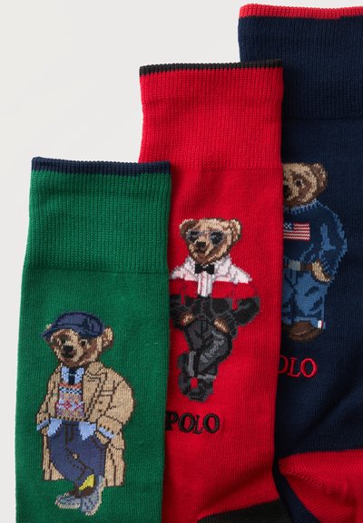 Trois chaussettes colorées avec des ours habillés de tenues élégantes sur fond vert, rouge et bleu marine avec la marque Polo.