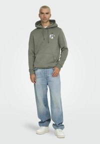 Groene hoodie met een voorzak en capuchon, voorzien van een kleurrafie op de borst. Gecombineerd met lichtblauwe wijde jeans en witte sneakers.