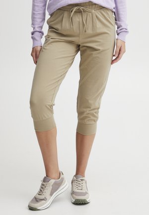 Beige avkuttede bukser med elastisk midjebånd og snøring, med smalnende mansjetter. Brukt med lysegrå og beige joggesko.