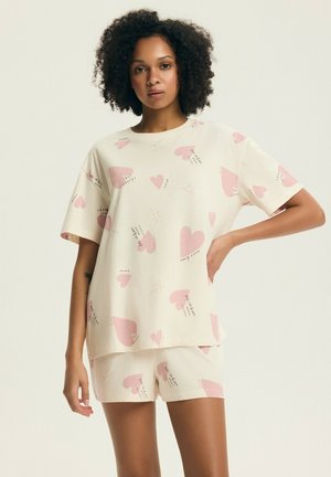Kobieta ubrana w kremowy zestaw piżamowy z różowymi serduszkami i małymi wzorami tekstowymi, stojąca z jedną ręką na biodrze na jednolitym tle.