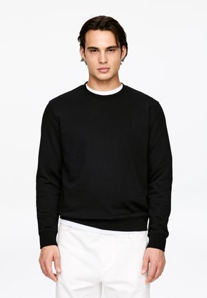 Hombre de cabello oscuro con sudadera negra de cuello redondo y pantalones blancos, de pie contra un fondo blanco liso.