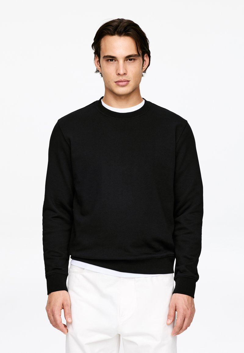 Homme aux cheveux foncés portant un sweat-shirt noir à col rond et un pantalon blanc, debout devant un fond blanc uni.