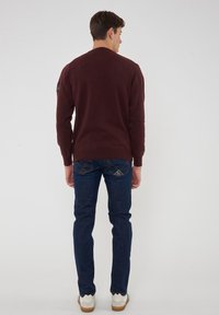 Maglione bordeaux a maniche lunghe con scollatura rotonda, abbinato a jeans slim-fit blu scuro. Scarpe bianche con bordi scallop, dettagli minimi.