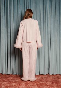 Žena s dlhými vlasmi otočená k modrým závesom, oblečená v textúrovanom svetloružovom saka a spodných častiach oblečenia s volánovými lemmi na rukávoch a členkoch.