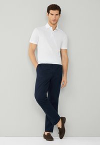 Polo shirt bianco a maniche corte, pantaloni tapered blu navy e scarpe slip-on in camoscio marrone. Linee pulite, design minimal e dettagli strutturati sulle scarpe.