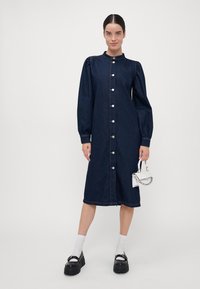 Denim obleka v temno modri barvi z dolgimi napihnjenimi rokavi, gumbi spredaj in visokim ovratnikom. Ima raven silhueto in rahle šive.