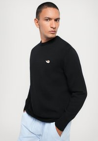 Svart strikket genser med rund hals. Har et lite, brodert logo på brystet. Kombinert med lyseblå-stripete bukser.