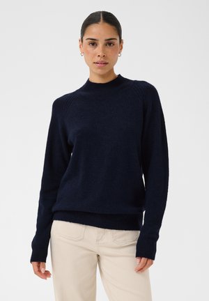 Femme aux cheveux foncés attachés, portant un pull bleu marine et un pantalon beige clair sur un fond clair uni.