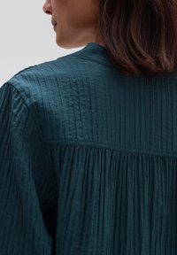 Türaqua-farbenes, strukturiertes Bluse mit vertikalen Falten, die einen weichen Kragen und ein gerafftes Rückendesign aufweist. Leichtes, atmungsaktives Material.