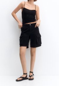 Mustat cargo-shorts sivutaskuilla, yhdistettyinä tiukkaan mustaan toppiin. Päällä litteät mustat sandaalit. Sileä kangas ja rento istuvuus.