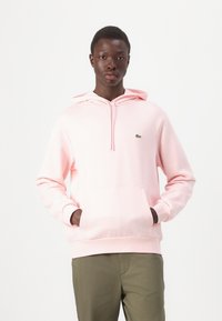 Sudadera de color rosa claro hecha de una tela suave, con capucha con cordón, bolsillo canguro y un pequeño logo verde en el lado izquierdo del pecho. Combinada con pantalones caqui.