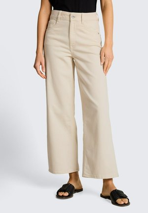 Personne portant un pantalon beige ample à jambes larges avec un bouton à l'avant et des sandales noires à enfiler, debout devant un fond clair et uni.