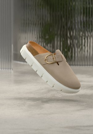 Μπεζ παπούτσι mule χωρίς κορδόνια με λευκή χοντρή σόλα, φελτίνη πάτο και διακοσμητική χρυσή αγκράφα σε θολό ουδέτερο φόντο.