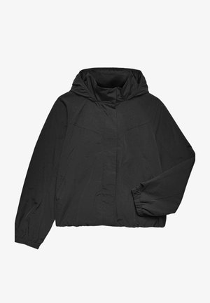 Ung kvinde iført en sort jakke med elastiske manchetter og et cremefarvet skørt med sorte polkaprikker mod en neutral baggrund.