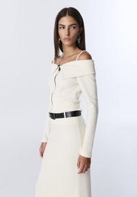 Top bianco a spalle scoperte in maglia con dettaglio in zip nera, abbinato a una gonna bianca coordinata e una cintura nera, con maniche lunghe.