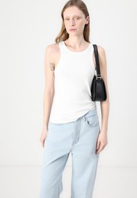 Hvit ribbet tank topp kombinert med lyseblå vid bukser. Modellen holder en liten svart veske med skulderrem. Minimalistisk design.