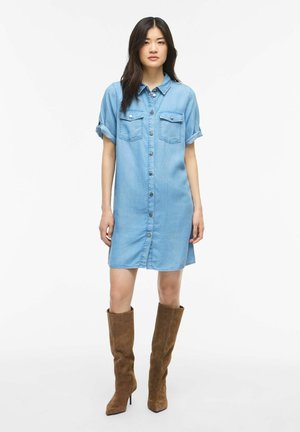 Femeie purtând o rochie din denim bleu deschis cu nasturi, mâneci suflecate și cizme maro de piele întoarsă până la genunchi, stând pe un fundal alb simplu.