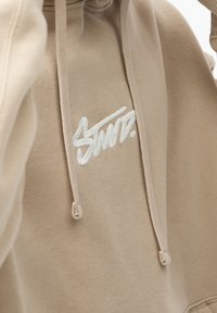 Beige Kapuzenpullover aus weichem Stoff, ausgestattet mit einer Front-Kängurutasche, einem Kordelzug an der Kapuze und einem gestickten weißen Logodetail.