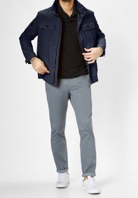 Laivaston kevyt takki, jossa on kaksi rintataskua, yhdistetty mustaan polo-paitaan ja vaaleanharmaisiin slim-fit-chinoihin. Valkoiset lenkkarit täydentävät asua.