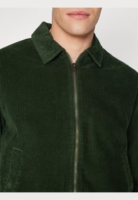 Donkergroene corduroy jas met een ritsluiting en puntige kraag, voorzien van verticale ribbelstructuur en zijzakken.