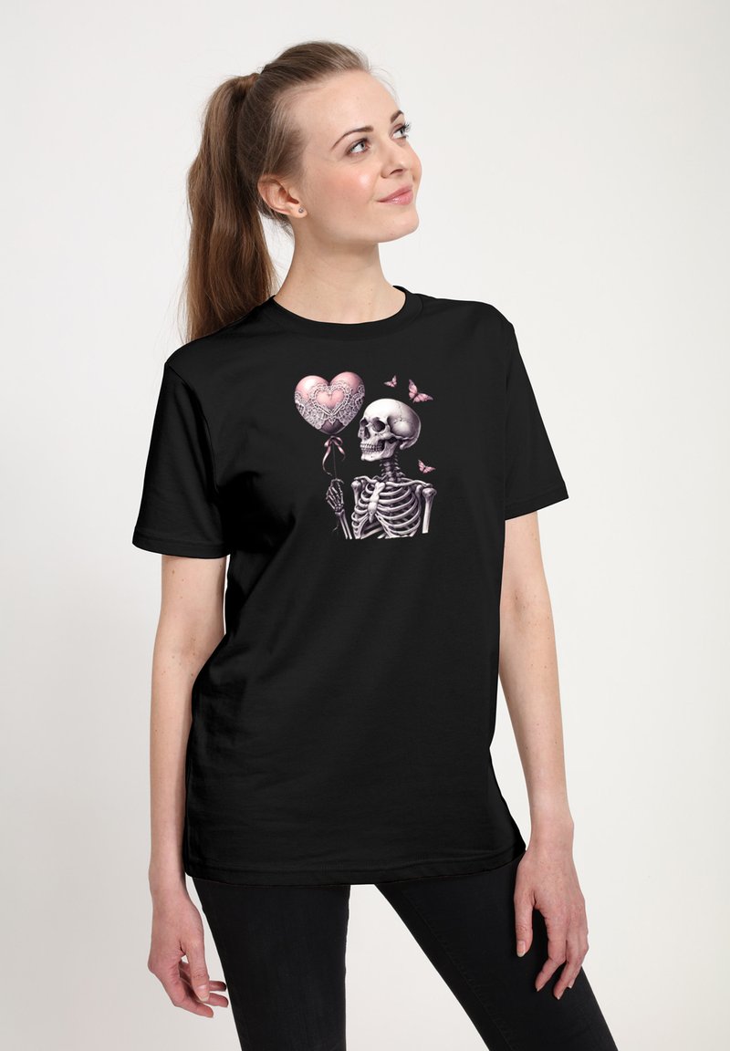 Woman in black t-shirt with a skeleton holding a pink heart-shaped balloon and pink butterflies, standing and smiling up to the side.