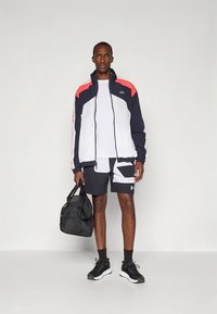 Meeste spordiriietus, mis koosneb tumesinisest, punasest ja valgest lukuga joped, valgest t-särgist, mustade mustriliste lühikeste pükstega ja mustadest tossudest. Käes must spordikott.