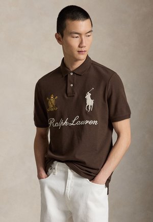 Jeune homme portant un polo Ralph Lauren marron avec logo et emblème, pantalon blanc, mains dans les poches, regardant sur le côté sur un fond neutre.