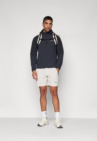 Black zip-up jacket with a hood, beige accents, white shorts, and light sneakers. Standing with a backpack, facing forward.