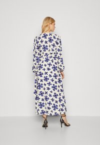 Robe longue blanche avec un motif floral en bleu et violet. Présente des manches longues, une taille cintrée et une silhouette fluide. Portée avec des talons noirs.