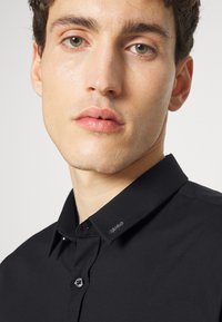 Camisa negra de manga larga hecha de una tela suave, con un cuello abotonado, un sutil detalle de logo en el cuello y un diseño ajustado.
