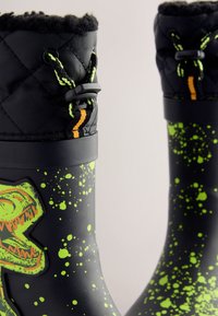 Stivali di gomma neri con motivo a schizzi verdi, decorati con una grafica di un dinosauro verde, collo imbottito in tessuto nero trapuntato e coulisse regolabile.