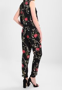 Svart blommig jumpsuit med ärmlös design, livfulla röda blomtryck, gröna detaljer och tapered ben med vändkanter.