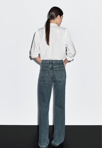 Hvid skjorte med lange ærmer og pufærmer, kombineret med blå jeans i høj talje med let slidte detaljer. Set bagfra fremhæves den skræddersyede pasform og lommer.