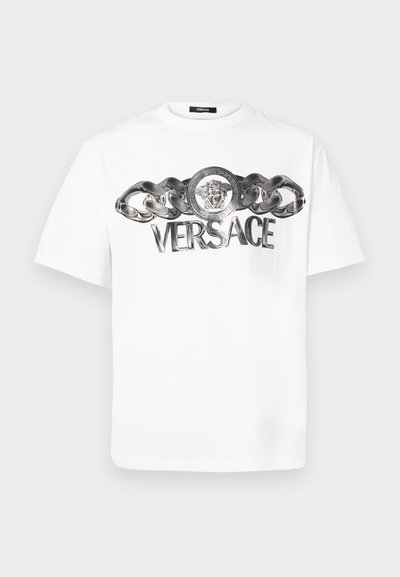 Valge puuvillane T-särk, millel on metallist kettide disain ja ümmargune embleem lõviga, millel on selgelt nähtav "VERSACE" logo.