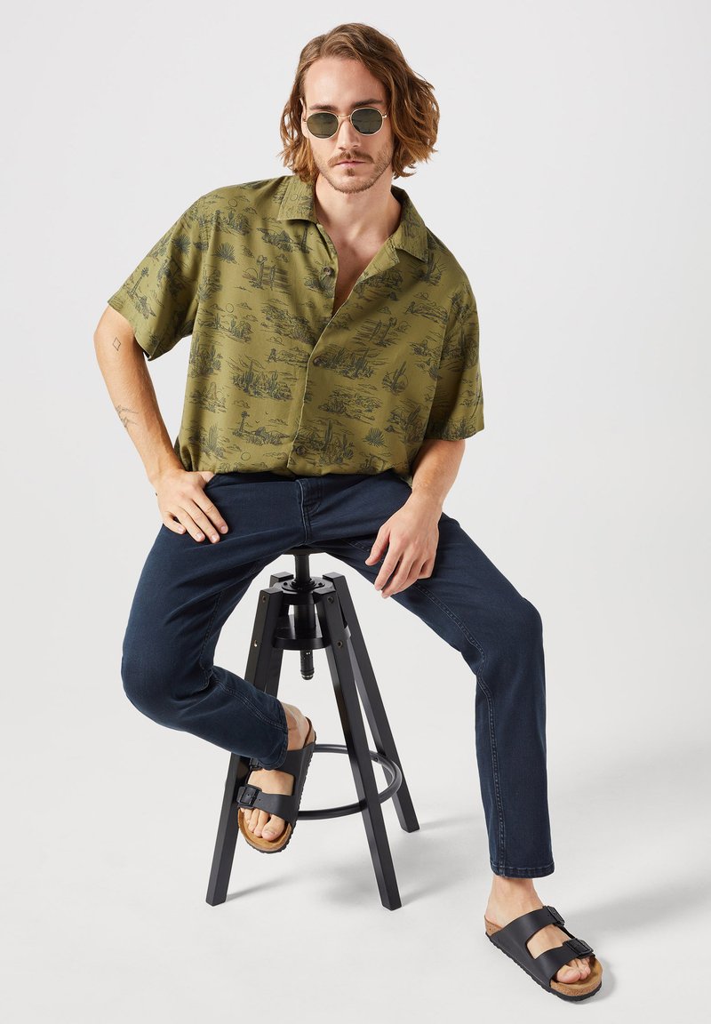 Camisa de manga corta verde oliva con estampado paisaje negro, combinada con jeans azul oscuro y sandalias negras. Modelo sentado en un taburete negro.