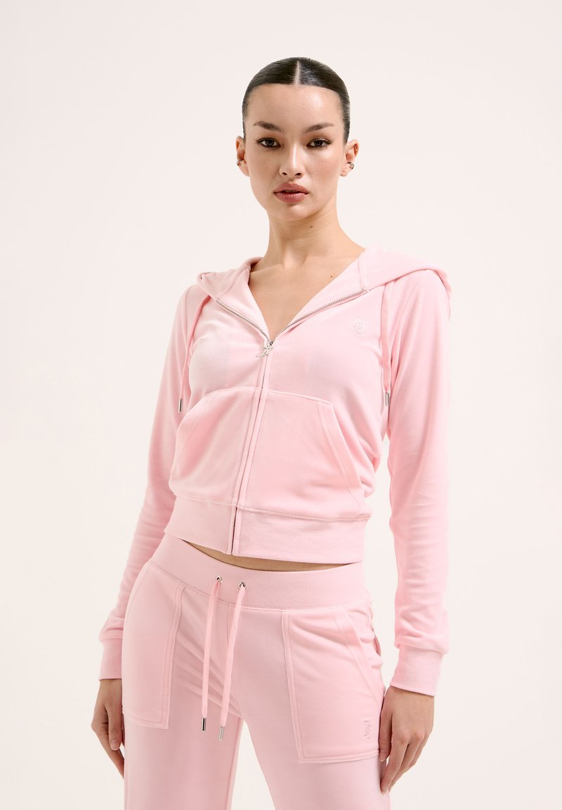 Ensemble de sport rose comprenant un sweat à capuche zippé court avec une poche frontale et un pantalon de jogging taille haute assorti avec cordon de serrage et poches latérales.