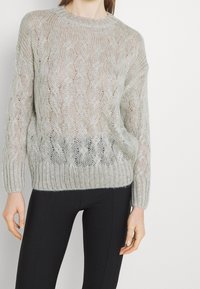 Pull en tricot gris clair avec un motif en câble texturé, coupe ample et poignets côtelés. Idéal pour superposer sur des vêtements ajustés.