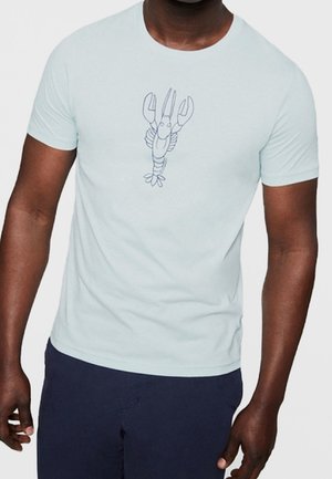 Mand iført lyseblå t-shirt med en enkel hummer-skitse placeret midt på brystet, kombineret med mørkeblå bukser.