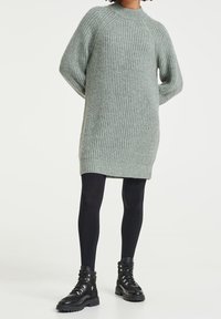Pletené šaty v světle šedé barvě s žebrovanou texturou, s vysokým krkem a oversized rukávy, spárované s černými legínami a armádními botami.