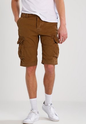 Mand iført brune cargoshorts, hvide sokker, hvide sneakers og en hvid t-shirt, stående foran en ensfarvet baggrund.