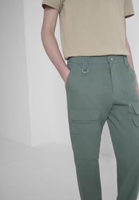 Groene cargobroek met een elastische tailleband, meerdere zakken en een D-ring detail. Gecombineerd met een beige T-shirt. Gladde textuur.